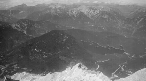 Das schwarz-weiß- Schrägluftbild zeigt eine Gebirgslandschaft. Im Vordergrund einen hellgrauen Berg, im Mittelgrund einen See und dunklere Berge und im Hintergrund wieder hellere Bergspitzen.